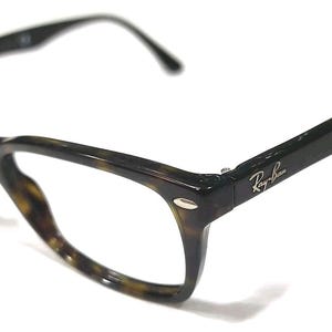 May include: Tortoiseshell-patterned eyeglasses with a dark brown and black frame. The temples feature the Ray-Ban logo in silver lettering near the hinges. These glasses have a classic rectangular shape with rounded edges and thin, curved arms.
