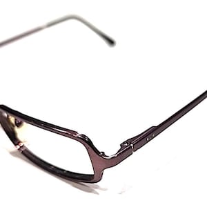 Puede incluir: Un par de gafas con montura morada oscura y varillas finas. Las lentes son transparentes y la montura tiene un diseño angular único. Las gafas están sobre un fondo blanco.