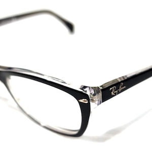 May include: Ray-Ban eyeglasses featuring a two-tone frame design with black upper rims and clear lower rims. The temples are black with a silver-toned Ray-Ban logo on the side. Small silver-toned metal accents are visible at the outer corners.