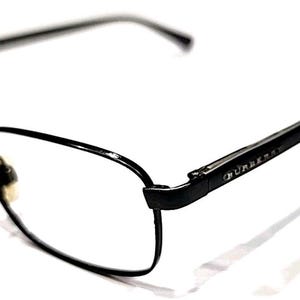 Puede incluir: Gafas rectangulares negras con una fina montura metálica. Las patillas presentan el nombre de la marca "Burberry" en una pequeña fuente plateada. Las lentes son transparentes y las gafas están sobre un fondo blanco.