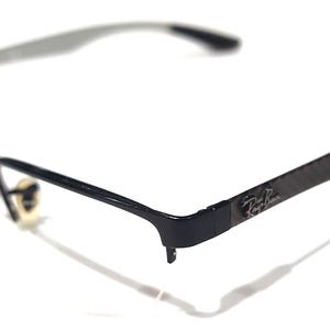 May include: Black eyeglasses featuring a thin metal frame and carbon fiber textured temples. The Ray-Ban logo is visible on the side of the temple. The glasses include adjustable nose pads and thin, lightweight arms designed for a secure fit.