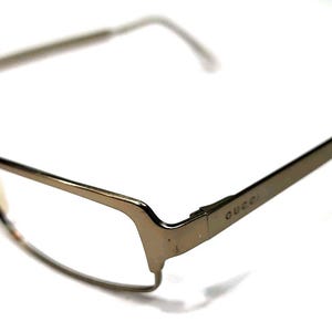 May include: A pair of semi-rimless eyeglasses with a metallic gold frame. The temples feature the Gucci brand name engraved on the side. The design includes thin metal arms and clear lenses, with a distinct bridge connecting the two frame sections.