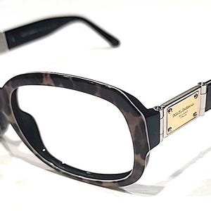 May include: Tortoiseshell-patterned eyeglasses with a rounded frame shape. The temples feature a gold-tone rectangular metal plaque engraved with the text Dolce & Gabbana Milano Italia, connected to black arms with a smooth, polished finish.