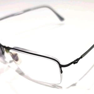 May include: A pair of rimless spectacles with a black metal frame. The lenses are clear, and the arms are thin and extend back to the ear pieces. The spectacles are set against a white background.