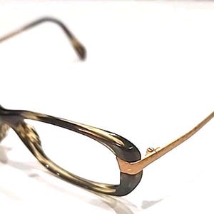 May include: Eyeglasses featuring a brown and tan tortoiseshell pattern on the oval-shaped frames. The temples are constructed from a thin, metallic gold-toned material, connecting to the front frames with a visible hinge mechanism on each side.