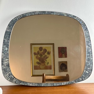 Könnte beinhalten: Ein rechteckiger Spiegel mit abgerundeten Ecken, umrahmt von einem Mosaik aus kleinen, grauen und weißen Fliesen. Der Spiegel reflektiert einen Raum mit Kunstwerken und einem Holzstuhl.