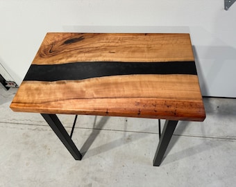 May include: A rectangular wooden table with a black epoxy resin river running through the center. The table has a natural wood grain finish and is supported by black metal legs. The table is approximately 30 inches long.