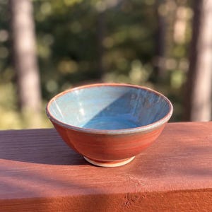 May include: A ceramic bowl with a smooth, glossy interior glazed in a light blue color. The exterior of the bowl features a warm, reddish-orange finish. The bowl has a circular base and a slightly flared rim, showcasing a two-tone design.