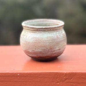 May include: A small, round, handmade ceramic pot with a wide opening. The pot features a light brown and cream color with subtle texture. The rim is slightly flared, and the base is a darker brown. The pot is likely used for decorative purposes.