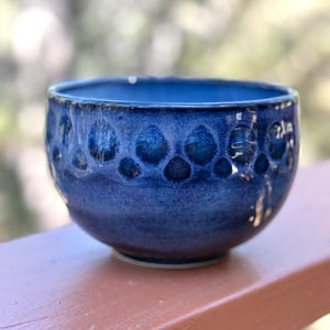 May include: A handmade, blue ceramic bowl with a textured, dimpled pattern around the exterior. The bowl has a glossy finish and a rounded shape, perfect for serving soups, stews, or other dishes. The bowl is sitting on a brown surface.