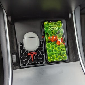 May include: A black phone charging pad with a red Tesla logo and a white wireless charging case for earbuds. A black iPhone with a green and orange fish screen saver is charging on the pad. The phone screen displays the time as 8:19.
