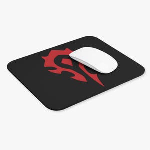 May include: Black rectangular mousepad with a red tribal design and a white wireless mouse. The mousepad features a stylized emblem. The mouse is positioned on the upper right side of the mousepad.