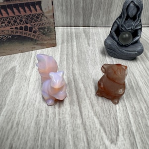 Puede incluir: Una colección de pequeñas figuras de piedra tallada. Las figuras incluyen una ardilla en tonos rosa y morado, un oso pardo y una estatua gris de una mujer sentada sosteniendo una esfera oscura. Una postal con la Torre Eiffel está en el fondo.