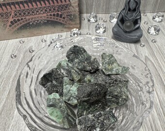 Puede incluir: Un cuenco de vidrio transparente lleno de piedras preciosas verdes oscuras y negras sin pulir. El cuenco está sobre una superficie de madera gris.