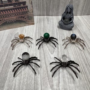 May include: Five decorative spider figurines with crystal orbs as bodies and metal legs. The spiders vary in orb color, including clear, green, and amber. A small statue and a print of the Eiffel Tower are in the background.