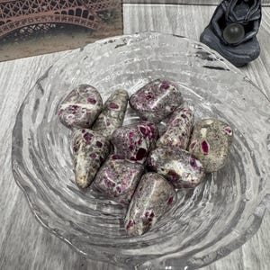 May include: A clear, textured glass bowl filled with tumbled stones. The stones are a mix of gray and white with flecks of deep red. A small statue and a print of the Eiffel Tower are in the background.