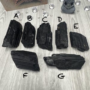 Puede incluir: Siete cristales de turmalina negra de diferentes tama&ntilde;os y formas. Los cristales est&aacute;n dispuestos sobre una superficie de madera gris claro.