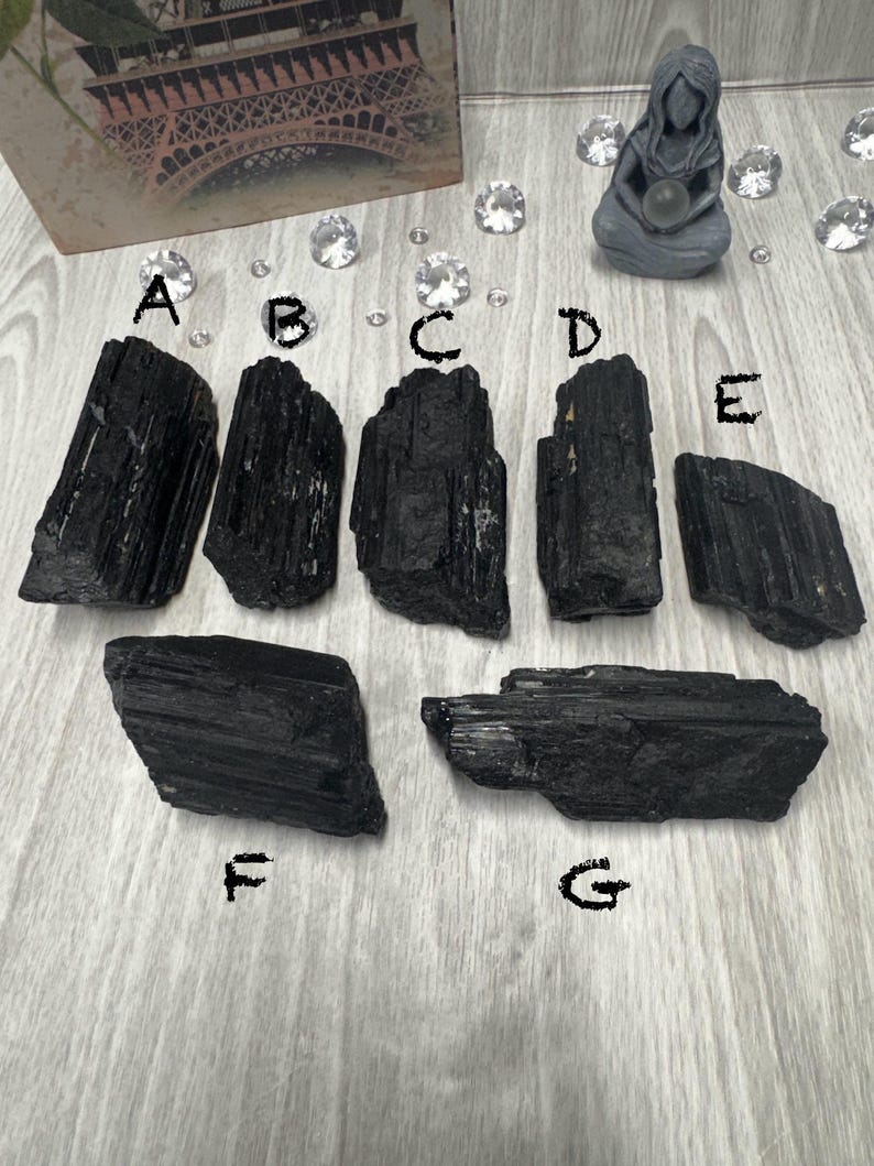 Puede incluir: Siete cristales de turmalina negra de diferentes tama&ntilde;os y formas. Los cristales est&aacute;n dispuestos sobre una superficie de madera gris claro.