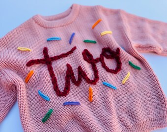 May include: Pink knitted sweater with the word "two" embroidered in burgundy yarn. The sweater is decorated with colorful sprinkles.
