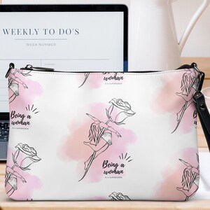 Puede incluir: Clutch blanco con una correa de muñeca negra. El diseño presenta una mujer sosteniendo una rosa, con acentos de acuarela rosa. El texto "Being a woman is a superpower" está impreso. Un portátil y un jarrón con flores rosas están en el fondo.