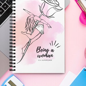 K&ouml;nnte beinhalten: Ein Spiralnotizbuch mit einer Schwarz-Wei&szlig;-Illustration einer Frau, die mit einer Rose tanzt. Der Text "Being a woman is a superpower" steht unter der Illustration. Der Hintergrund ist eine rosa Aquarellwaschung.