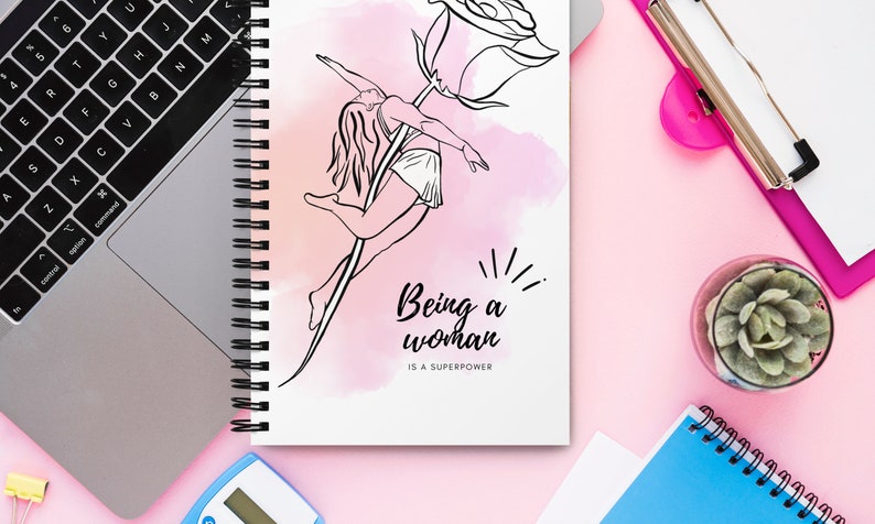 K&ouml;nnte beinhalten: Ein Spiralnotizbuch mit einer Schwarz-Wei&szlig;-Illustration einer Frau, die mit einer Rose tanzt. Der Text "Being a woman is a superpower" steht unter der Illustration. Der Hintergrund ist eine rosa Aquarellwaschung.