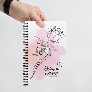 K&ouml;nnte beinhalten: Spiralgebundenes Notizbuch mit einer Schwarz-Wei&szlig;-Illustration einer Frau, die auf einem Rosenstiel tanzt. Das Design enth&auml;lt den Text "Being a woman is a superpower" &uuml;ber einem rosa Aquarellhintergrund.