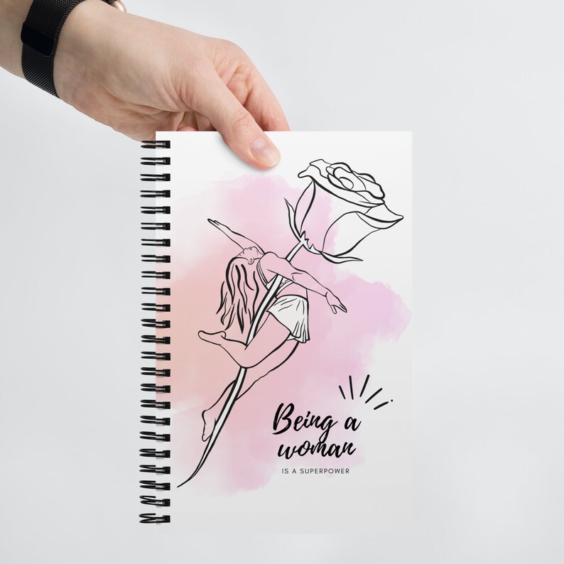 K&ouml;nnte beinhalten: Spiralgebundenes Notizbuch mit einer Schwarz-Wei&szlig;-Illustration einer Frau, die auf einem Rosenstiel tanzt. Das Design enth&auml;lt den Text "Being a woman is a superpower" &uuml;ber einem rosa Aquarellhintergrund.