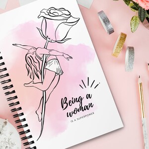 K&ouml;nnte beinhalten: Ein Spiralnotizbuch mit einer Schwarz-Wei&szlig;-Strichzeichnung einer Frau, die auf einem Rosenstiel tanzt. Der Text "Being a woman is a superpower" befindet sich unter der Zeichnung. Der Hintergrund ist rosa mit dekorativen Elementen.