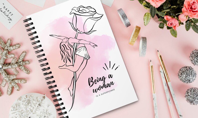 K&ouml;nnte beinhalten: Ein Spiralnotizbuch mit einer Schwarz-Wei&szlig;-Strichzeichnung einer Frau, die auf einem Rosenstiel tanzt. Der Text "Being a woman is a superpower" befindet sich unter der Zeichnung. Der Hintergrund ist rosa mit dekorativen Elementen.