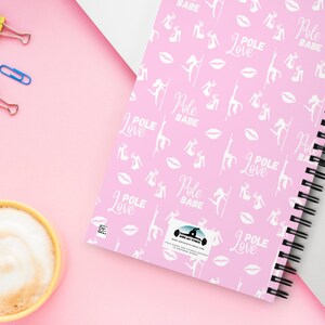 K&ouml;nnte beinhalten: Ein pinkfarbenes Notizbuch mit Spiralbindung, das ein Muster aus wei&szlig;en Pole-Dance-Figuren, Lippen und den W&ouml;rtern "Pole Love" und "Pole Babe" aufweist. Das Notizbuch liegt neben einem Laptop mit bunter Tastaturabdeckung, einer gelben Kaffeetasse und B&uuml;roklammern.