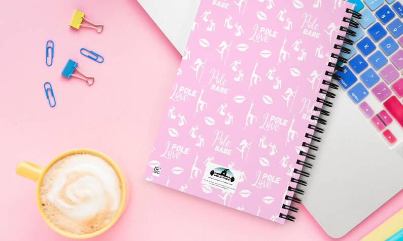 K&ouml;nnte beinhalten: Ein pinkfarbenes Notizbuch mit Spiralbindung, das ein Muster aus wei&szlig;en Pole-Dance-Figuren, Lippen und den W&ouml;rtern "Pole Love" und "Pole Babe" aufweist. Das Notizbuch liegt neben einem Laptop mit bunter Tastaturabdeckung, einer gelben Kaffeetasse und B&uuml;roklammern.