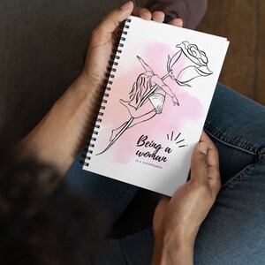 K&ouml;nnte beinhalten: Spiralgebundenes Notizbuch mit einer Schwarz-Wei&szlig;-Strichzeichnung einer Frau und einer Rose. Das Design ist vor einem rosa Aquarellhintergrund platziert. Der Text "Being a woman is a superpower" steht darunter.