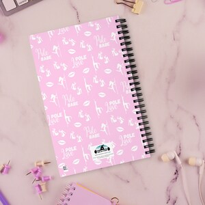K&ouml;nnte beinhalten: Rosa Spiralnotizbuch mit einem sich wiederholenden Muster aus wei&szlig;en Pole-Dance-Figuren, Lippen und den Worten "Pole Babe" und "Pole Love". Das Notizbuch ist von B&uuml;robedarf und einer Tasse Tee umgeben.