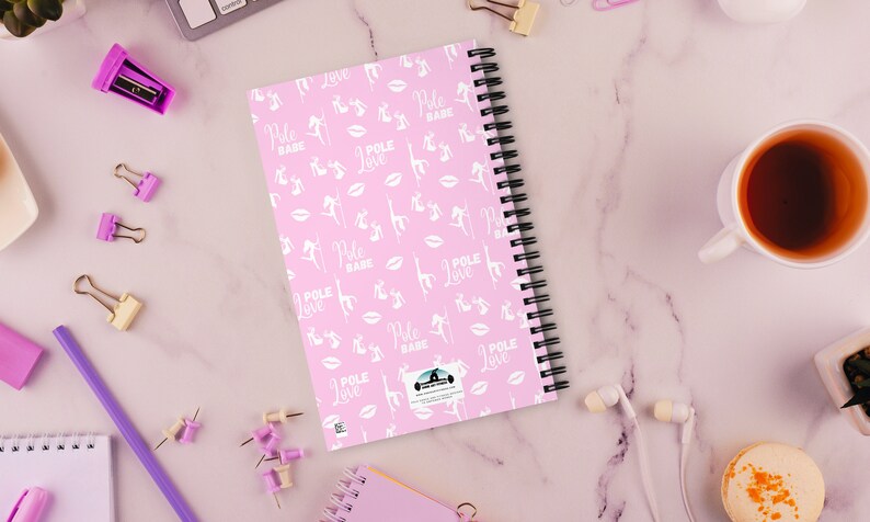 K&ouml;nnte beinhalten: Rosa Spiralnotizbuch mit einem sich wiederholenden Muster aus wei&szlig;en Pole-Dance-Figuren, Lippen und den Worten "Pole Babe" und "Pole Love". Das Notizbuch ist von B&uuml;robedarf und einer Tasse Tee umgeben.