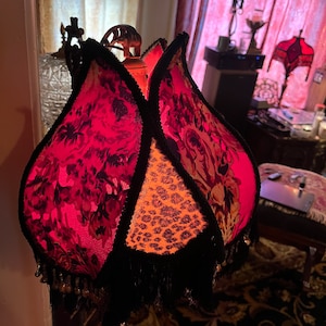 May include: A decorative lampshade with a unique, tulip-like design. The shade features panels of red floral fabric, leopard print, and black trim with beaded fringe. The lamp has a brass-colored metal base and is lit from within.