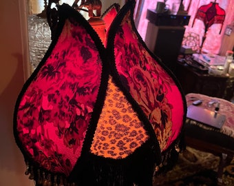 May include: A decorative lampshade with a unique, tulip-like design. The shade features panels of red floral fabric, leopard print, and black trim with beaded fringe. The lamp has a brass-colored metal base and is lit from within.
