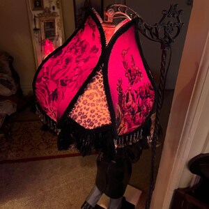 May include: A unique floor lamp with a floral patterned shade in shades of pink and black. The shade is trimmed with black fringe and a leopard print fabric. The lamp has a black metal frame and a checkered base.