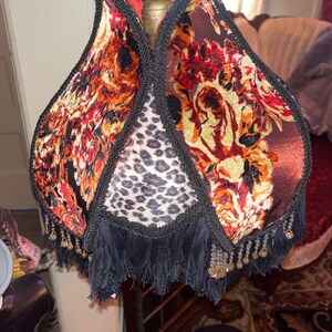 May include: A decorative lamp with a unique shade. The shade features a floral pattern in shades of red, orange, and brown, with a leopard print panel. The shade is trimmed with black fringe and beading.