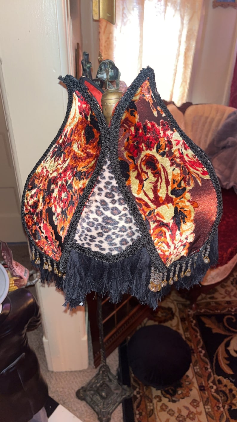 May include: A decorative lamp with a unique shade. The shade features a floral pattern in shades of red, orange, and brown, with a leopard print panel. The shade is trimmed with black fringe and beading.