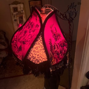 May include: A decorative floor lamp with a vibrant pink lampshade featuring a floral pattern and black fringe. The lamp has a black metal frame and a leopard print center. The lamp is a unique home decor item.