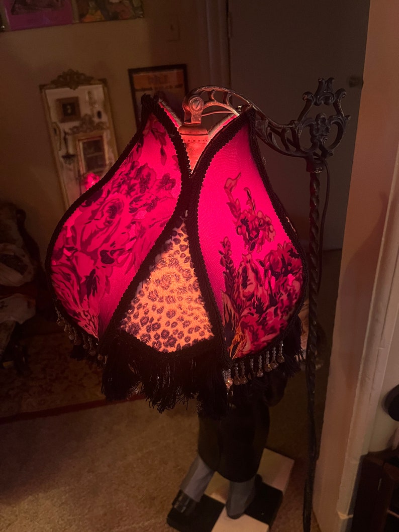 May include: A decorative floor lamp with a vibrant pink lampshade featuring a floral pattern and black fringe. The lamp has a black metal frame and a leopard print center. The lamp is a unique home decor item.
