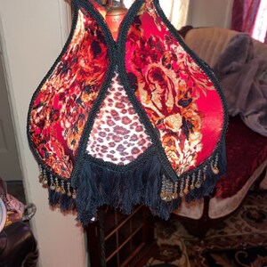 May include: A decorative floor lamp with a unique shade. The shade features a red floral pattern with black trim and fringe. The center panel has a leopard print design. The lamp has a dark metal base.