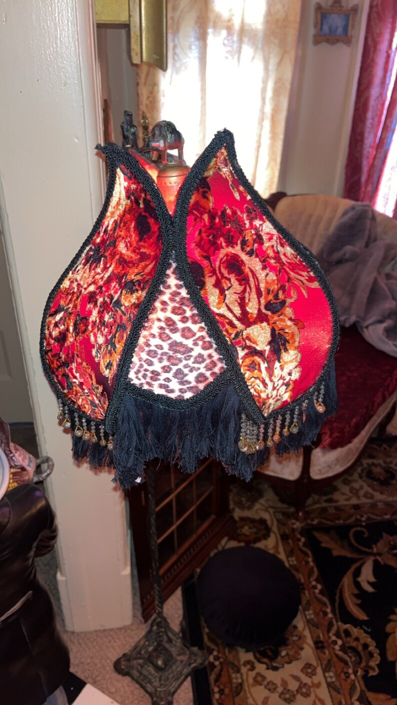 May include: A decorative floor lamp with a unique shade. The shade features a red floral pattern with black trim and fringe. The center panel has a leopard print design. The lamp has a dark metal base.