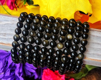 Könnte beinhalten: Ein Stapel schwarzer Edelstein-Armbänder mit goldenen Sprenkeln. Die Armbänder bestehen aus runden Perlen und sind auf einer Holzoberfläche gestapelt.