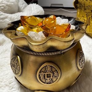 Könnte beinhalten: Ein goldfarbener Metalltopf mit einem dekorativen Design und chinesischen Schriftzeichen. Der Topf ist mit weißen Steinen und drei goldfarbenen Barren gefüllt.