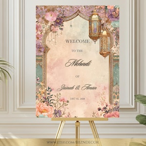 May include: A decorative welcome sign for a Mehndi ceremony, featuring floral and lantern motifs. The sign includes the text "Welcome to the Mehndi of Zainab & Aman" and the date "DEC 02, 2029". The sign is displayed on a gold easel.