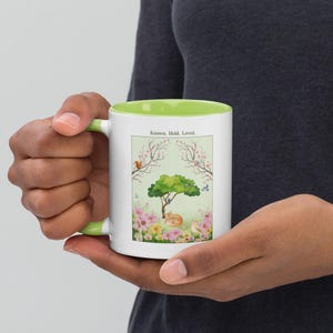 Könnte beinhalten: Weißer Keramikbecher mit grünem Innenraum und Griff. Der Becher zeigt eine Naturszene mit einem Baum, Blumen und einer schlafenden Katze. Der Text "Known. Held. Loved." ist oben auf dem Design aufgedruckt.