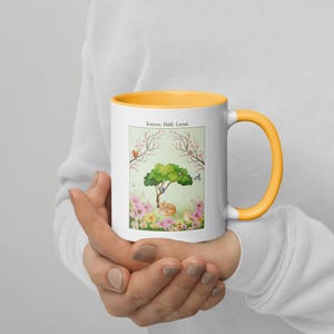 Könnte beinhalten: Weißer Keramikbecher mit gelbem Innenraum und Henkel. Der Becher zeigt eine skurrile Illustration eines schlafenden Fuchses unter einem Baum, umgeben von Blumen und Schmetterlingen. Der Text "Known. Held. Loved." ist oben aufgedruckt.
