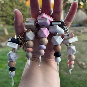 May include: Several keyrings with book-shaped charms are displayed. The keyrings feature a variety of beads in neutral tones, geometric shapes, and small book charms in white, pink, and black. Each keyring has a silver ring.